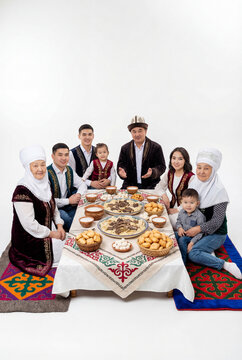 Large Central Asian family in traditional clothes having dinner at dastarkhan table. Kazakh family gathering with national dishes like beshbarmak, baursak, and kumis on white background.