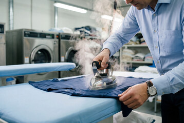 Naklejka premium Male laundry worker ironing blue shirt with steam in professional laundromat