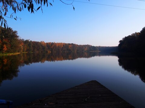 Autumn morning at Pusty Staw in the Stogi, Gdańsk, Poland. Calm lake water reflects colorful trees and clear blue sky, creating a serene natural landscape. 