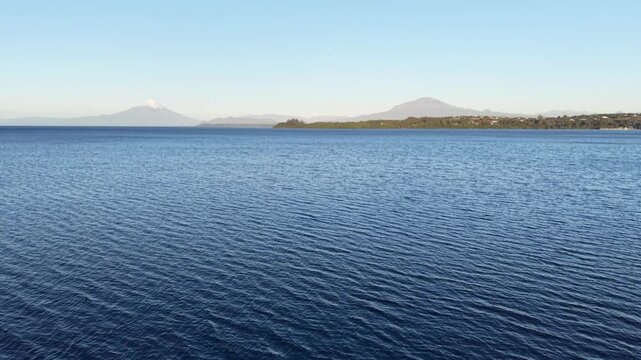 Cinematic aerial moving forward from the Puerto Varas shoreline over Lake Llanquihue, revealing the snow-capped Osorno and Calbuco volcanoes in the Chilean Lake District
