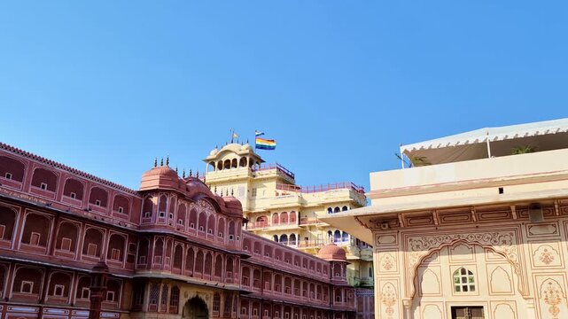 The Chandra Mahal rises behind the pink walls of the City Palace, Jaipur. The five-colored flag of the Jaipur royal family flies atop the historic seven-story structure against a bright blue sky.