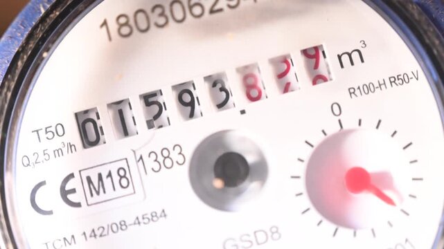 Close-up video of a domestic water meter in operation. Red indicator needles and small rotating dials show real time water consumption. Concept of utility measurement, resource management
