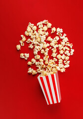 Popcorn exploding from striped bucket on red background. Creative cinema snack concept.