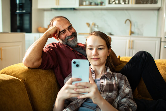 Father and teen daughter using smartphone on cozy sofa at home