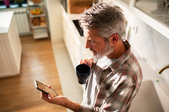 Mature man drinking coffee and using smartphone in home kitchen