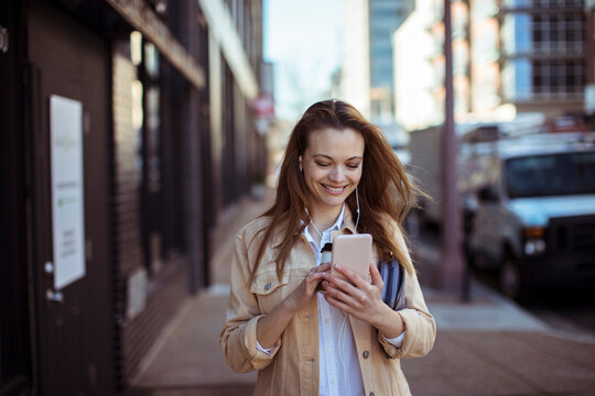 Smiling young woman with earphones using smartphone on city street