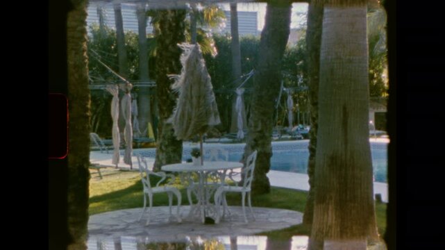 A static medium full shot of an empty pool patio with a white wrought iron table and two chairs near a folded sun umbrella with long tassels, blowing in the breeze at a resort on vintage super 8 film