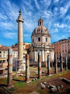  Ancient Roman Forum ruins with Trajan's Column and Santa Maria di Loreto church under a bright blue sky with clouds, Rome, Italy.
