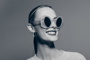Portrait of a smiling Caucasian woman with light skin wearing round sunglasses and hoop earrings. Minimalist studio grey background emphasizes her stylish and confident look. © SHOTPRIME STUDIO