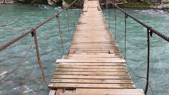 First-person view crossing a rickety and dangerous wooden suspension bridge. The shaky structure spans a fast-flowing turquoise river