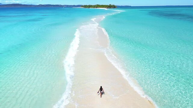 Aerial drone view of a woman walking along a narrow white sandbar between shallow turquoise waters, with a small island visible in the distance in Madagascar