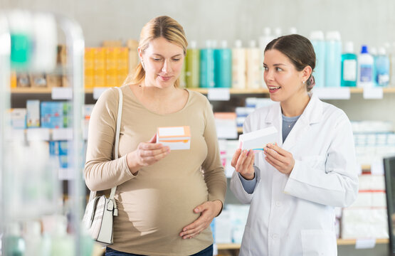 Pregnant woman choose pills of iodide and folic acid in pharmacy consulting with pharmacist, examine packaging. Drug store staff offer supplements to customer