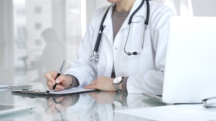 Female doctor in white coat with stethoscope writing patient notes on clipboard, completing medical...