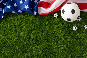 A soccer ball and mini balls rest on green grass beside an American flag evoking USA patriotism and summer holiday sports and Independence celebration