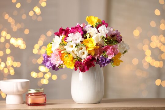 Beautiful freesia flowers in vase and candle on wooden table indoors. Bokeh effect