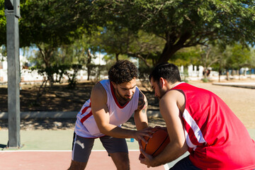 Obraz premium Two young men playing basketball in a park, dribbling the ball during a competitive outdoor game on a sunny day