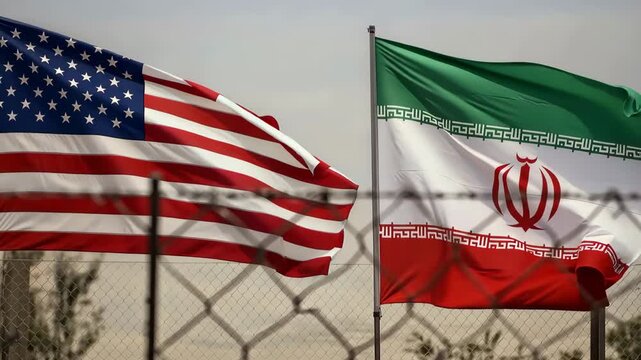 United States and Iran flags waving behind a chain link fence. Geopolitical rivalry and diplomatic tension between nations. Global crisis and international relations conflict concept.