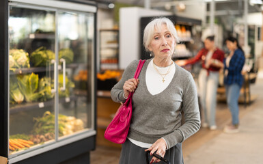 Fototapeta premium Visiting store elderly woman is looking at something on shelf of showcase, client buy goods in background