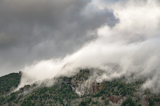 Dramatic clouds rolling over forested mountain ridges with exposed rock faces in moody atmospheric conditions, creating a mystical landscape scene in the Appalachian mountains