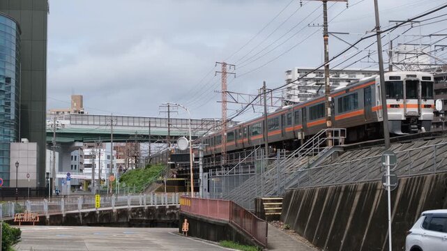 Commuter train passing through an urban railway corridor in Nagoya, Japan with overhead power lines, elevated tracks, surrounding city infrastructure. Rail transport in a dense Japanese neighborhood