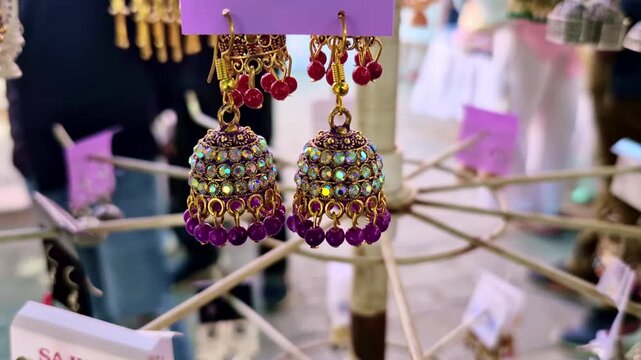 Sparkling purple and gold jhumka earrings with red accents hang on display. These handcrafted pieces at a Jaipur market showcase the vibrant jewelry traditions found near the Hawa Mahal.