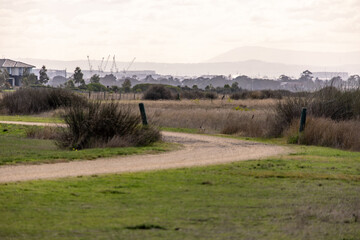 Fototapeta premium Natural grassland and walking trail in Cheetham Wetlands, Point Cook, Melbourne, Australia with distant suburban housing and soft atmospheric haze. Open coastal reserve near urban neighborhood