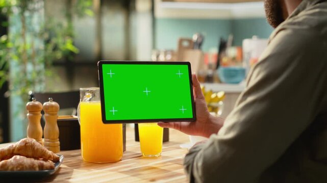Mockup chroma key on tablet screen in the kitchen at wooden table. Modern lifestyle with green screen, infographic elements and digital tools, teleworking and multitasking indoors.