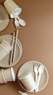 Eco-friendly disposable tableware arrangement with biodegradable paper plates, kraft paper cups, wooden cutlery, and paper straws on warm brown background.