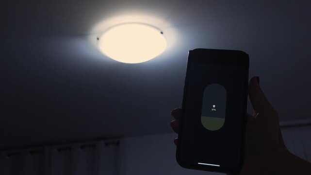 Hand holding smartphone with smart light control interface, adjusting brightness of ceiling light in a dimly lit room
