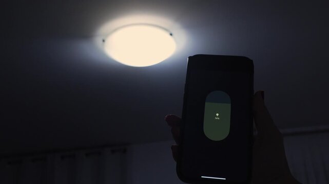 Hand holding smartphone adjusts brightness of ceiling light, showcasing gradual illumination change in a dimly lit room with curtains