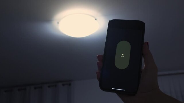 Hand holding smartphone with illuminated screen, adjusting light settings in a modern room with ceiling light fixture and curtains in the background