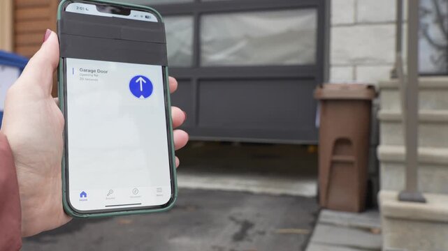 Hand holding smartphone displaying garage door control interface, showing progression of door opening in residential driveway setting