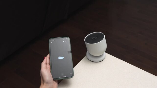 Hand holding smartphone displays app interface while interacting with smart speaker on a table in a modern living room setting