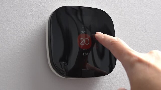 Hand adjusts smart thermostat display showing temperature settings in a modern home environment with a clean wall background