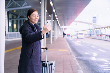 Fototapeta premium Asian female traveler with luggage taking selfie or video call at airport bus stop