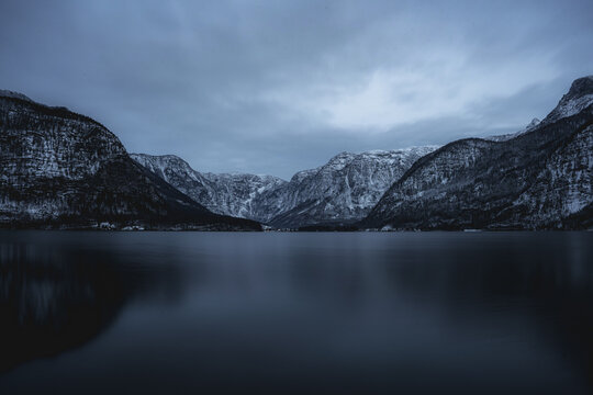 Moody winter landscape of a calm alpine lake surrounded by snow-covered mountains along the route between Kaprun, Austria and Munich, Germany. The still water reflects the dramatic alpine scenery 