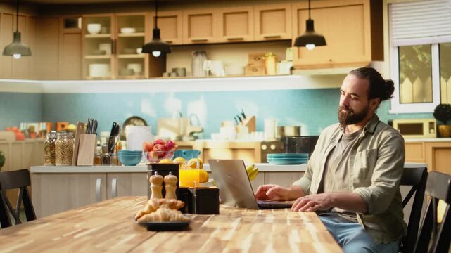 Adult man working from home in a cozy rustic kitchen, using laptop for daily tasks online. Freelancer lifestyle with remote work setup and natural light supporting productivity and multitasking.