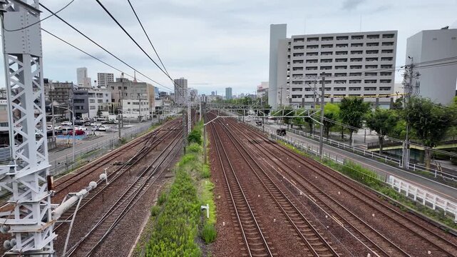 Commuter train traveling through a multi-track railway corridor in Nagoya, Japan with overhead power lines and dense urban buildings. Wide rail network cutting through a modern Japanese cityscape.