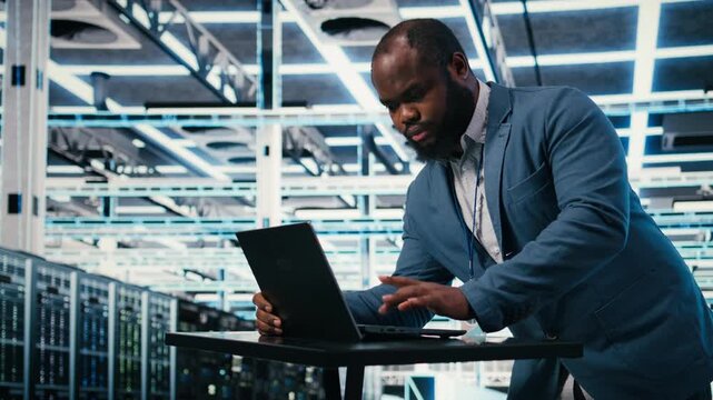 Programmer at server farm workplace desk using laptop, doing network management. Data center IT expert overseeing IT infrastructure supercomputers with software, typing on notebook keyboard, camera A