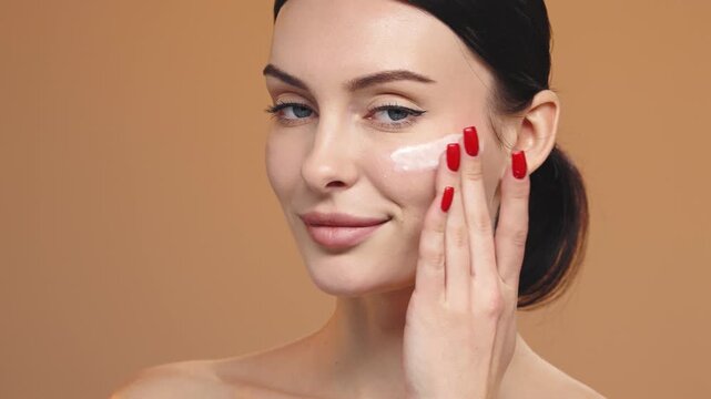 Smiling woman applying cosmetic serum on face isolated on studio background. Skincare and wellness concept.
