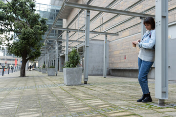 Asian woman leaning by post at plaza checking watch and holding coffee backpack visible, copy space © wavebreak3