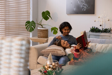 Diverse couple reclining on light-colored sofa at home, man reading red hardcover book