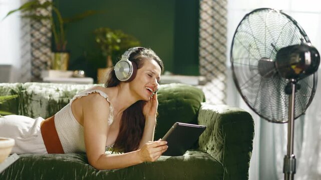 A smiling woman lies comfortably on a green sofa, engaging with a digital tablet while wearing headphones. A fan provides cooling, creating a perfect atmosphere for leisure and entertainment at home.