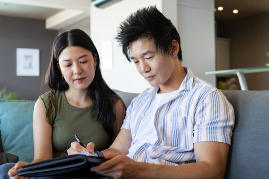 Asian couple sitting on gray sofa in living room reviewing black portfolio and signing with pen