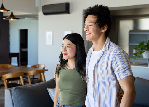 Diverse adult couple standing, smiling, looking right in open-plan home with gray sofa and pendants