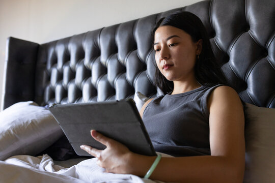 Asian woman reclining on bed by headboard in gray top holding tablet folio cover, copy space