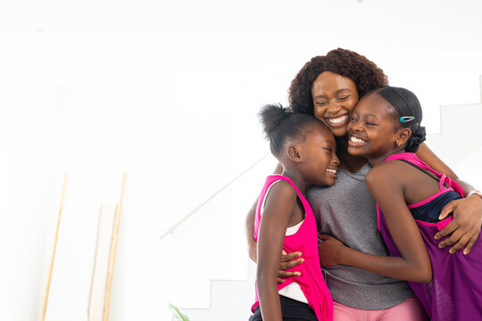 Hugging mother and girls wearing gray top, pink tanks at home, with staircase, boards, copy space