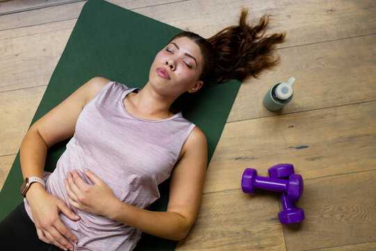 Resting woman wearing mauve tank, leggings on green mat at studio, with dumbbells, bottle, watch