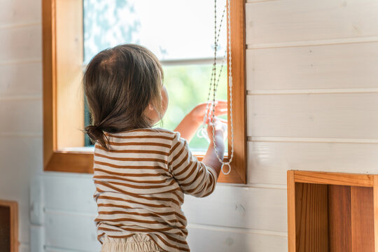 A mischievous girl's back as she plays by pulling on the cords of roller blinds and other blinds.