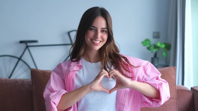 Head shot caucasian young woman sit on sofa look at camera put hands on chest make with fingers heart shape symbol of sincere feelings love. Volunteer donation, liberality loyalty sign concept&nbsp;
